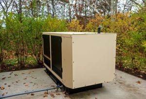 A residential standby generator sits on a concrete pad in a landscaped yard, with a fuel line connected and trees and shrubs surrounding it.