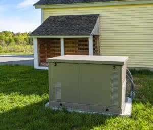 Outdoor home standby generator installed on a concrete pad beside a house and storage shed.