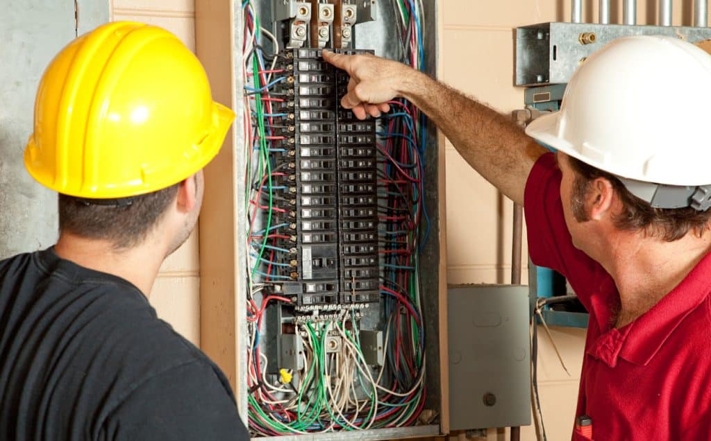 Two electricians in hard hats inspect an electrical panel filled with circuit breakers and wiring.